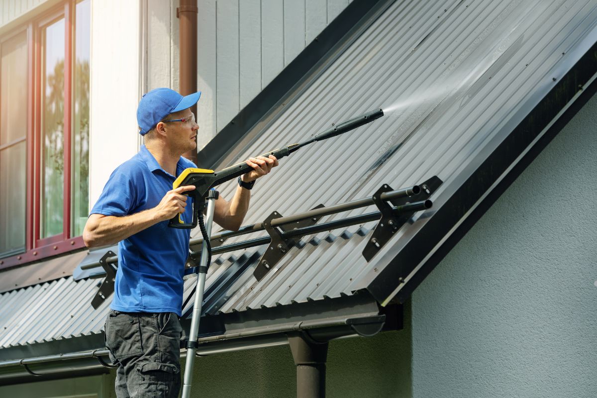 A person wearing a blue shirt cleans a metallic roof using a pressure washer, spraying water to remove dirt and grime.