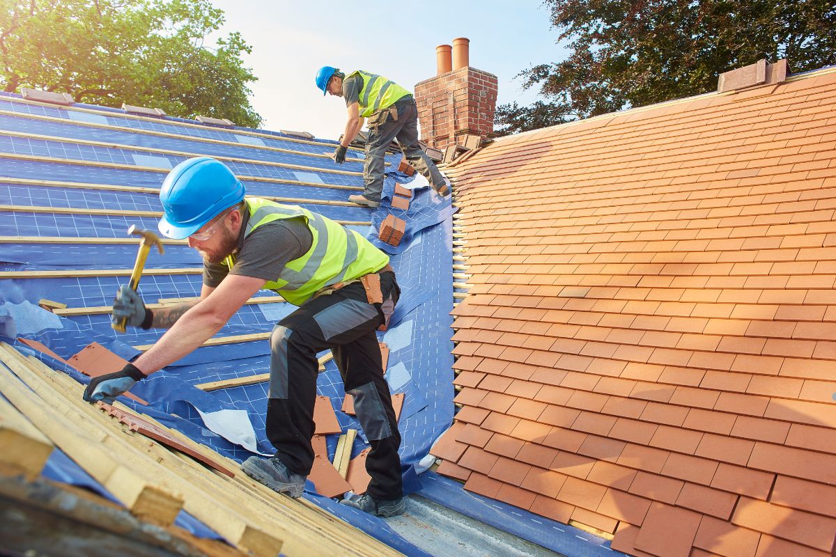 Two construction workers in hard hats and vests install roof tiles on a sloped roof, with tools and materials scattered around.