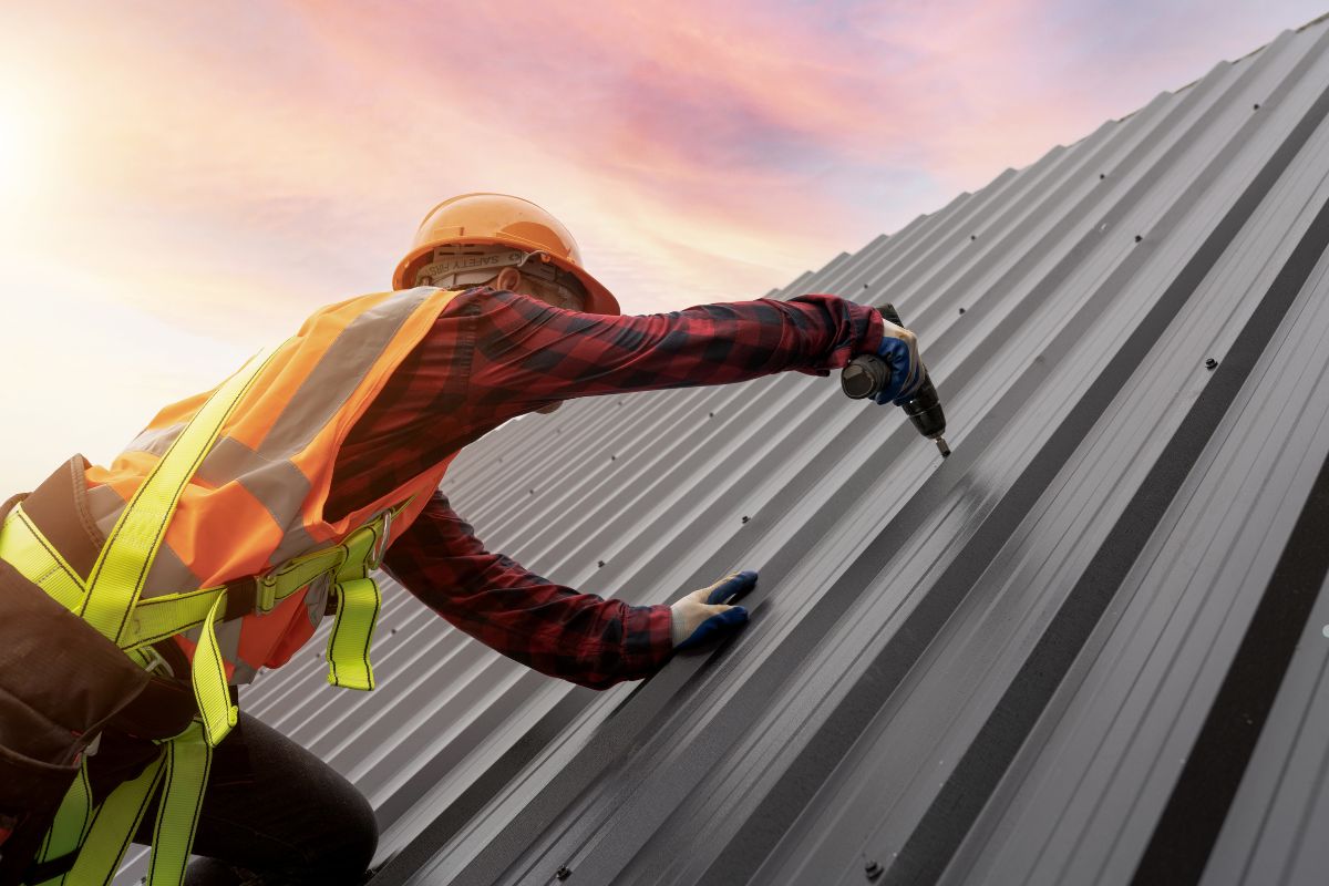 A construction worker in an orange safety vest and helmet drills into a metal roof at sunset, ensuring secure installation.