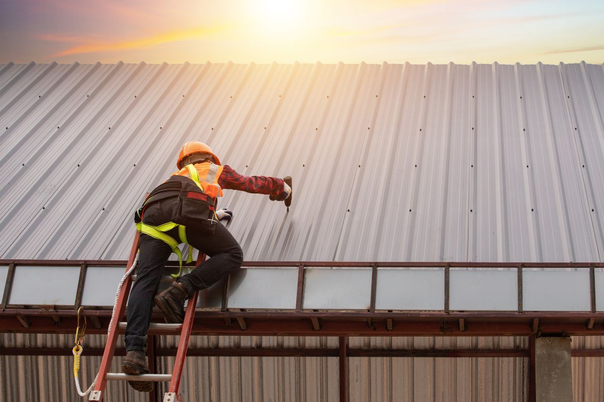 A worker in a safety harness climbs a ladder to repair or inspect a metal roof at sunset, wearing a hard hat and vibrant safety gear.