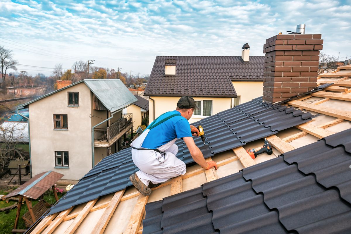 A person is installing black metal roofing tiles on a sloped roof, with houses and trees visible in the background under a blue sky.
