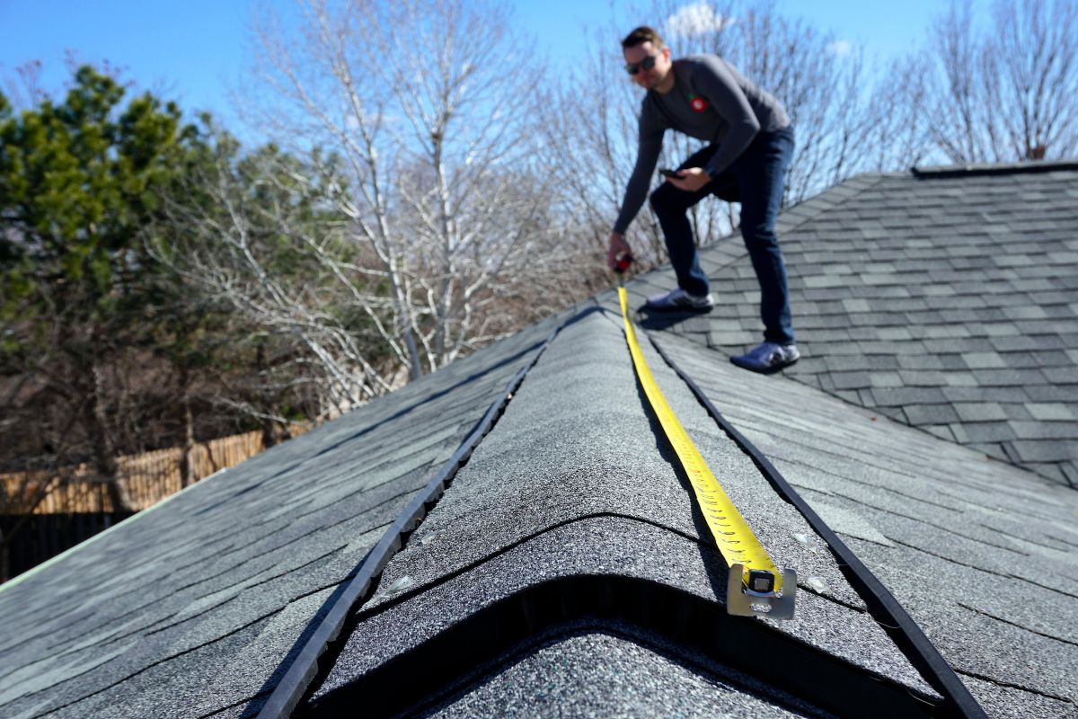 A person measures the ridge of a steep roof with a yellow tape measure, surrounded by trees and clear blue sky.