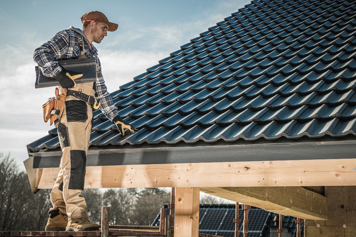 A man stands on a house roof, holding a tool, surveying the area around him.